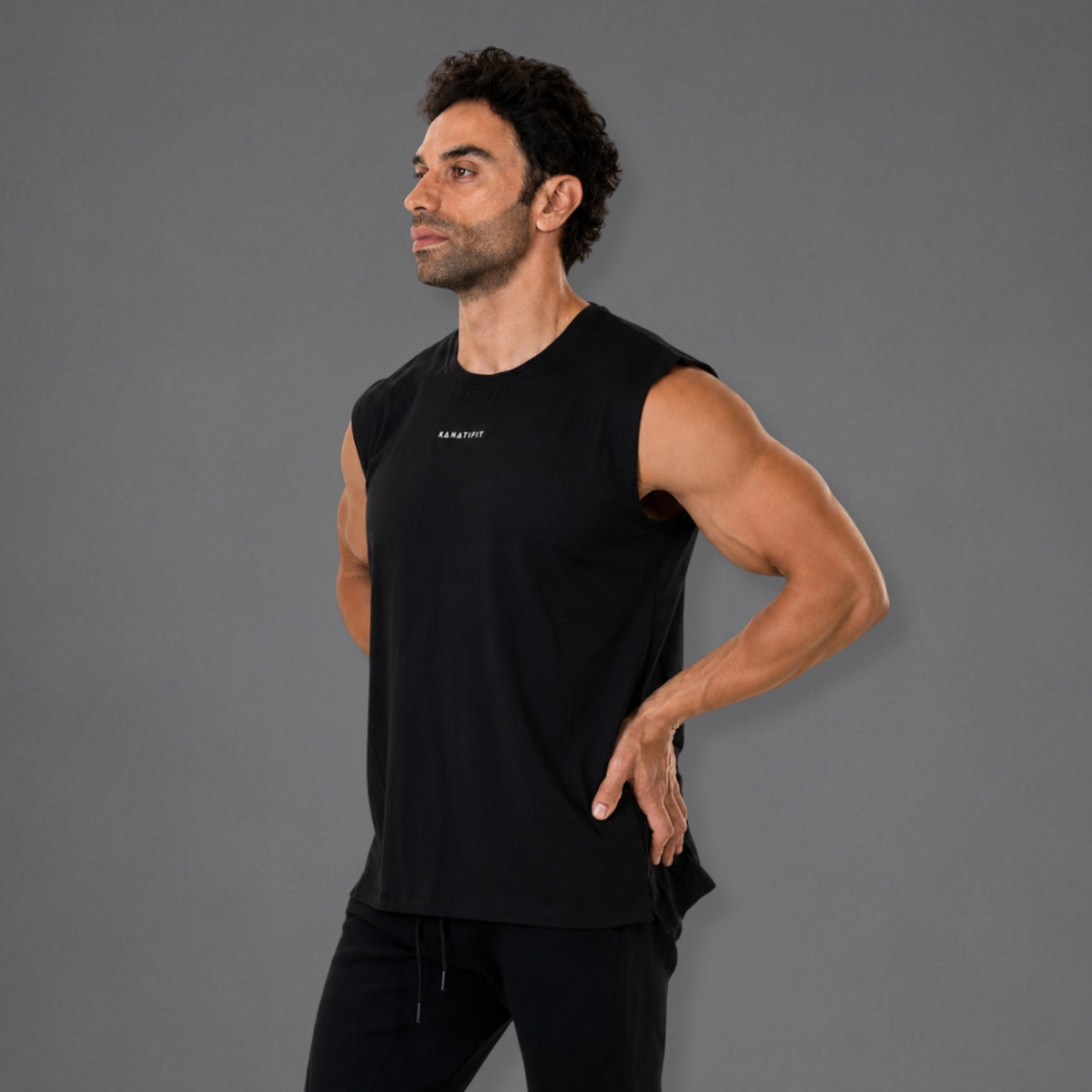 A man wearing a black sleeveless athletic tank top with a small white logo on the chest, paired with black pants, standing against a solid gray studio background.