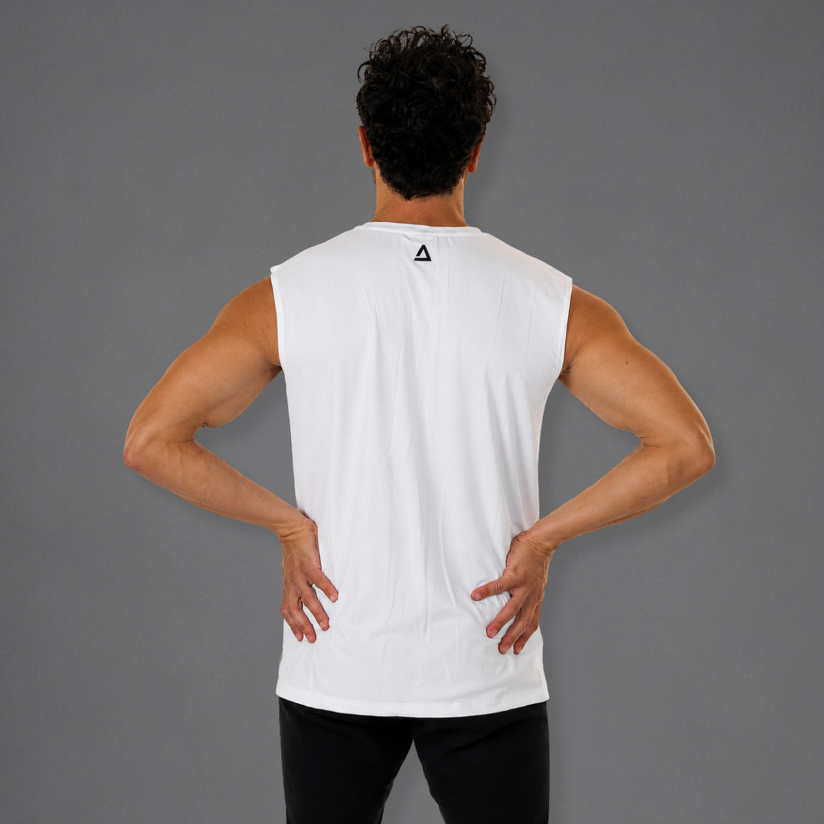 A man viewed from behind wearing a white sleeveless athletic tank top with a small black geometric logo on the upper back, paired with black pants, against a solid gray studio background.