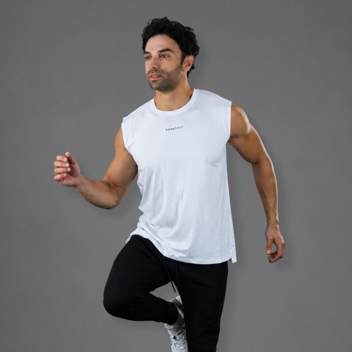 A man in a white sleeveless athletic tank top and black pants running against a solid gray studio background. The tank top has a small black logo on the chest that reads 'KANATIFIT'