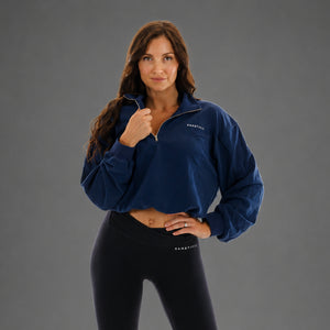 A woman modeling a navy blue cropped quarter-zip sweatshirt with white 'KANATIFIT' branding on the left chest, paired with black leggings, against a solid gray studio background.