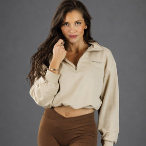A woman modeling a cropped beige half-zip pullover with a small 'KANATIFIT' logo on the chest, paired with brown leggings, against a solid gray studio background.