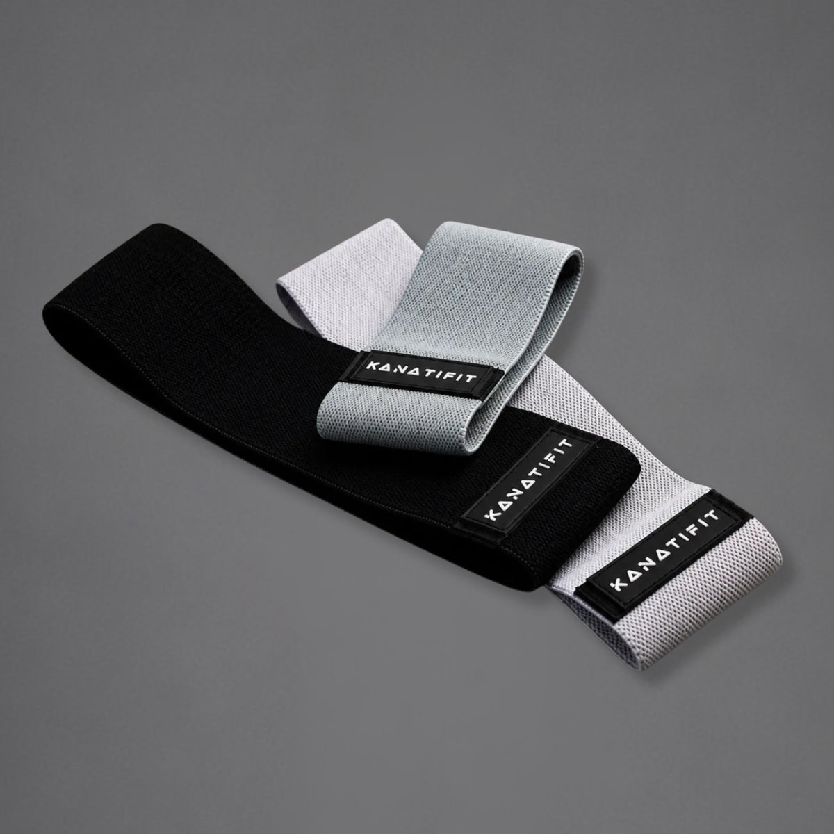 Three 'KANATIFIT' fitness resistance bands, one black, one grey and one light gray, arranged on a dark gray background.