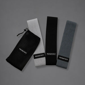 Three 'KANATIFIT' resistance bands in black, light gray, and dark gray, displayed next to a black mesh storage pouch on a solid gray background.