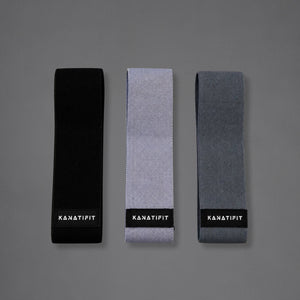 Three resistance bands flat on a gray background, each with a black label at the bottom reading 'KANATIFIT'. The straps are shown in three colorways: solid black, light heather gray, and medium charcoal gray.