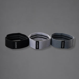 Three resistance bands in black, light gray, and dark gray, arranged side-by-side on a solid gray background. Each band features a black vertical label with the brand name 'KANATIFIT' in white.