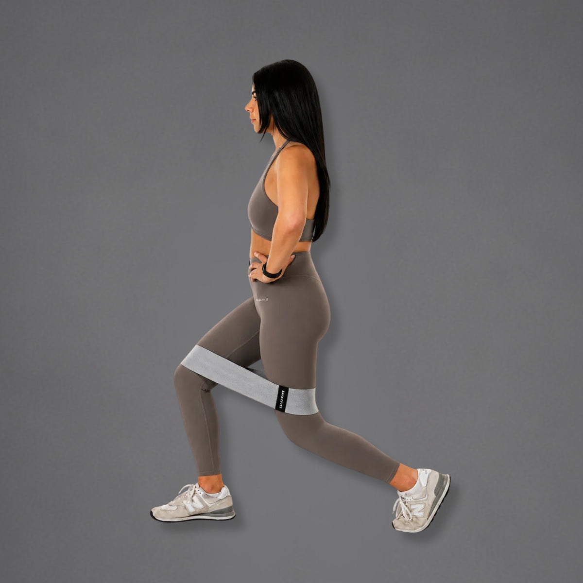 A woman in profile wearing a gray sports bra, taupe leggings, and white sneakers demonstrates a lunge position while wearing a light gray resistance band around her upper leg against a solid gray background.