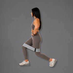 A woman in profile wearing a gray sports bra, taupe leggings, and white sneakers demonstrates a lunge position while wearing a light gray resistance band around her upper leg against a solid gray background.