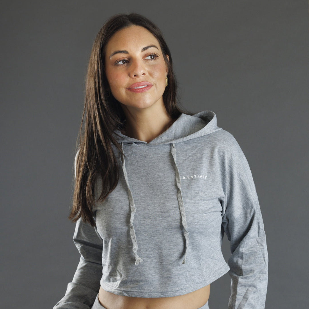 A woman wearing a grey cropped hoodie with long sleeves and drawstrings, photographed against a solid gray studio background.