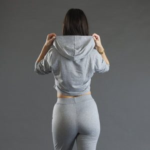A woman viewed from behind wearing a light gray heather cropped hoodie and matching high-waisted leggings against a solid dark gray studio background.