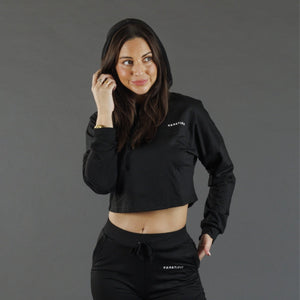 A woman modeling a black cropped hooded sweatshirt and matching black sweatpants against a solid gray studio background. The brand name 'KANATIFIT' is printed in white on the left chest of both garments.