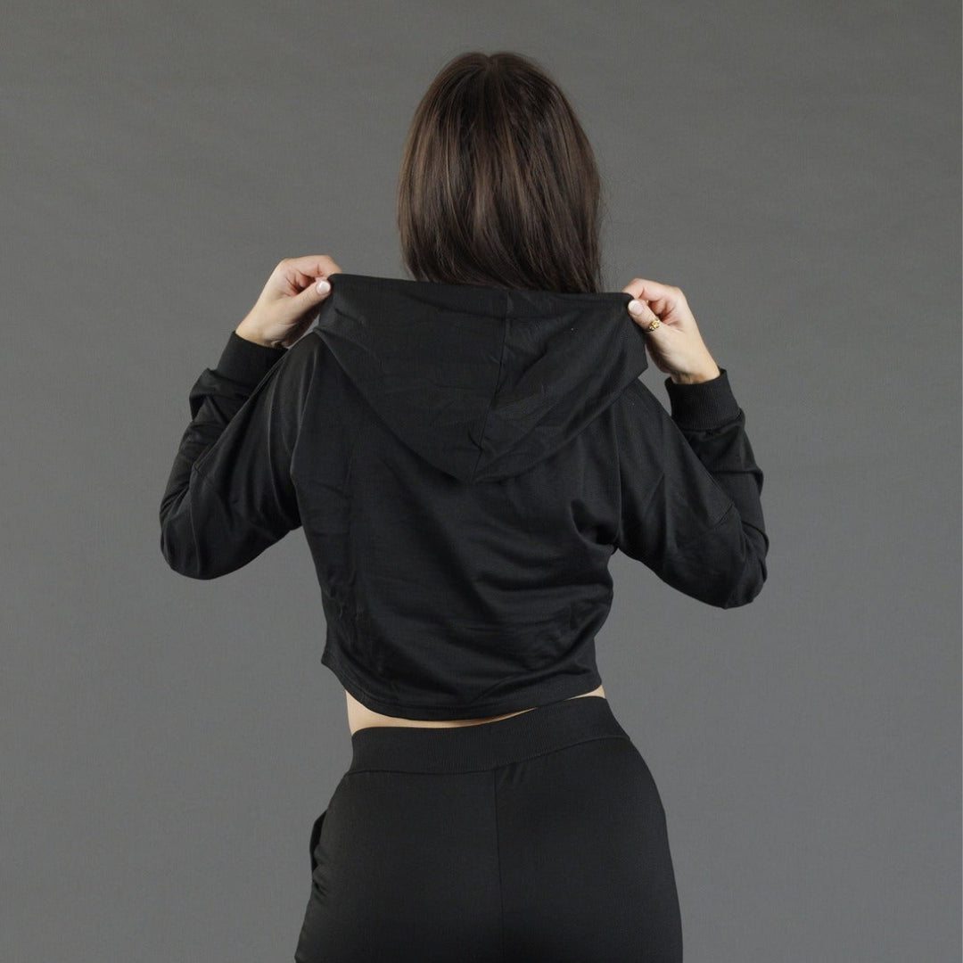A woman viewed from behind wearing a black cropped hooded sweatshirt and black pants, holding the hood up with both hands against a plain gray studio background.