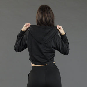 A woman viewed from behind wearing a black cropped hooded sweatshirt and black pants, holding the hood up with both hands against a plain gray studio background.