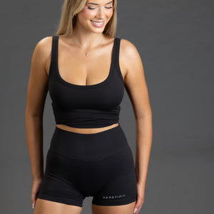 A woman modeling a black activewear set consisting of a scoop-neck crop top and high-waisted shorts with 'KANATIFIT' printed on the leg, set against a solid dark gray studio background.