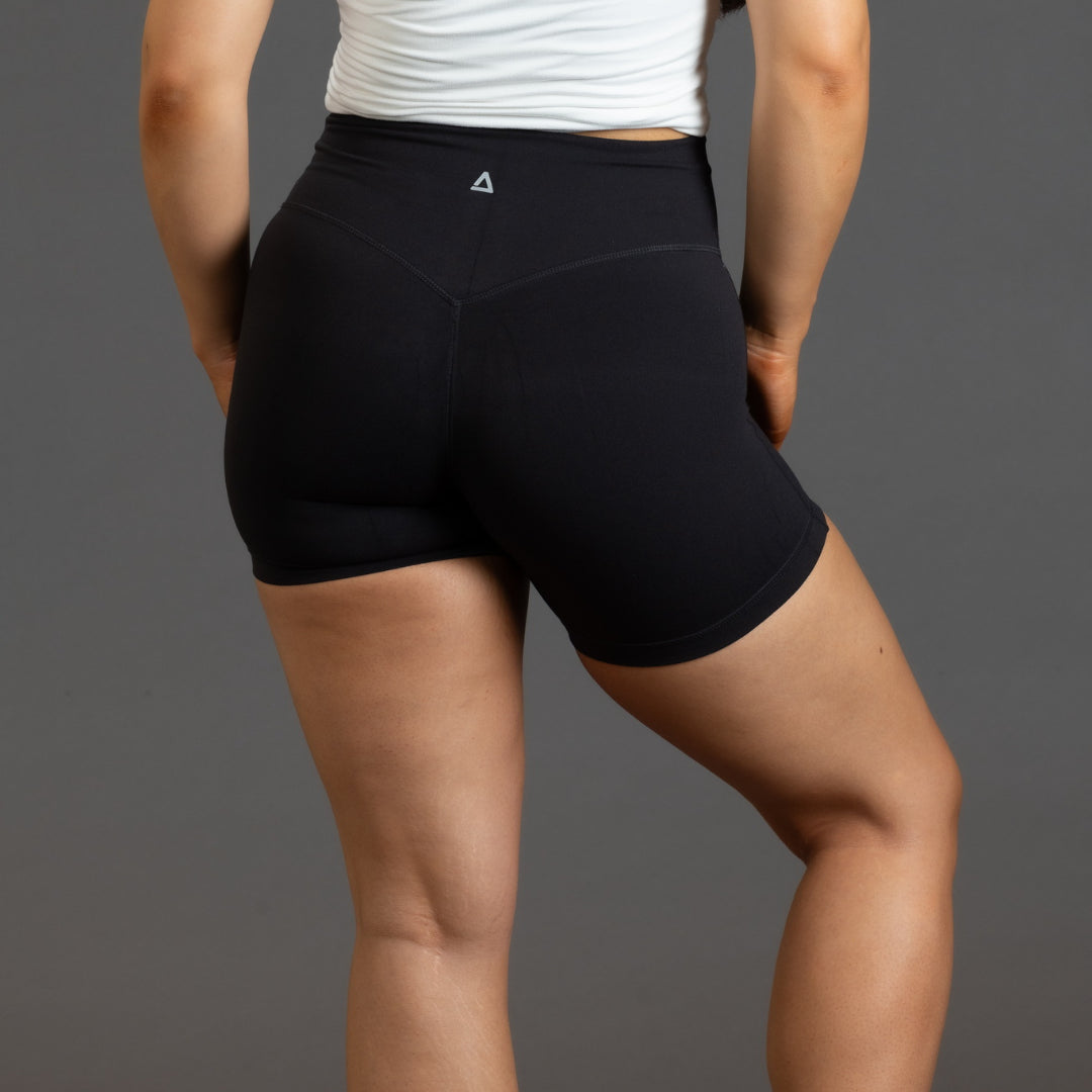 Rear view of a model wearing black high-waisted athletic shorts with a small white geometric 'KANATIFIT' logo on the back waistband, paired with a white tank top against a solid gray background.