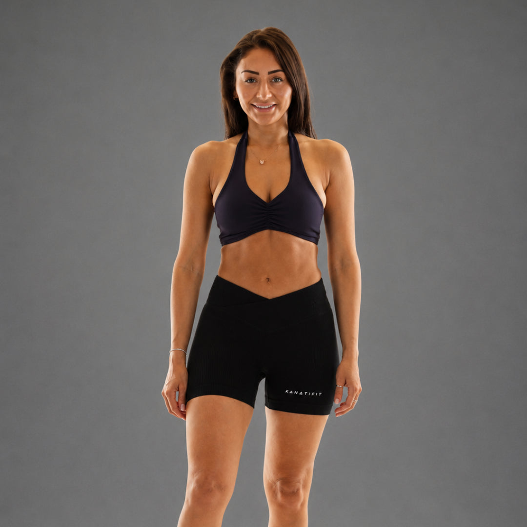 A woman modeling a dark navy blue sports bra and matching black athletic shorts against a solid gray studio background.