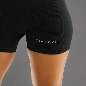 Close-up of a person wearing black ribbed athletic shorts with the brand name KANATIFIT printed in white on the side, against a plain gray background.