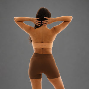 A woman shown from behind wearing a matching beige sports bra and brown high-waisted athletic shorts against a solid gray studio background.