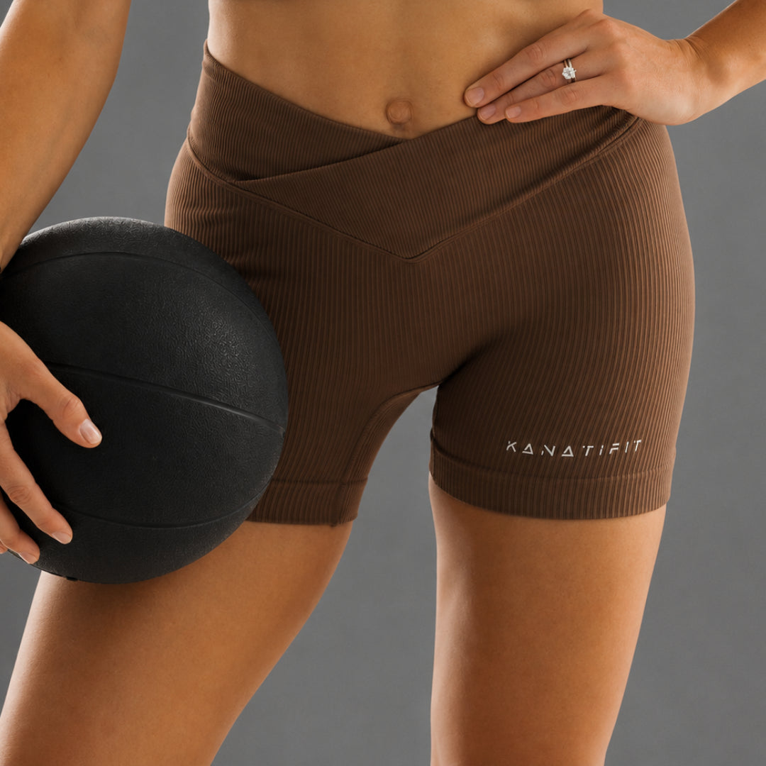 A close-up of a model wearing brown ribbed athletic shorts with the brand name 'KANATIFIT' printed in white on the lower right leg, holding a black medicine ball against a solid gray background.