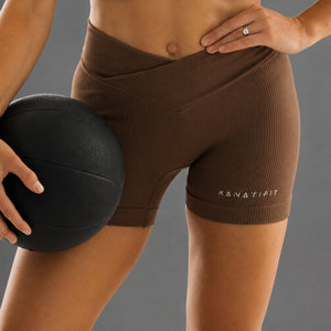 A close-up of a model wearing brown ribbed athletic shorts with the brand name 'KANATIFIT' printed in white on the lower right leg, holding a black medicine ball against a solid gray background.