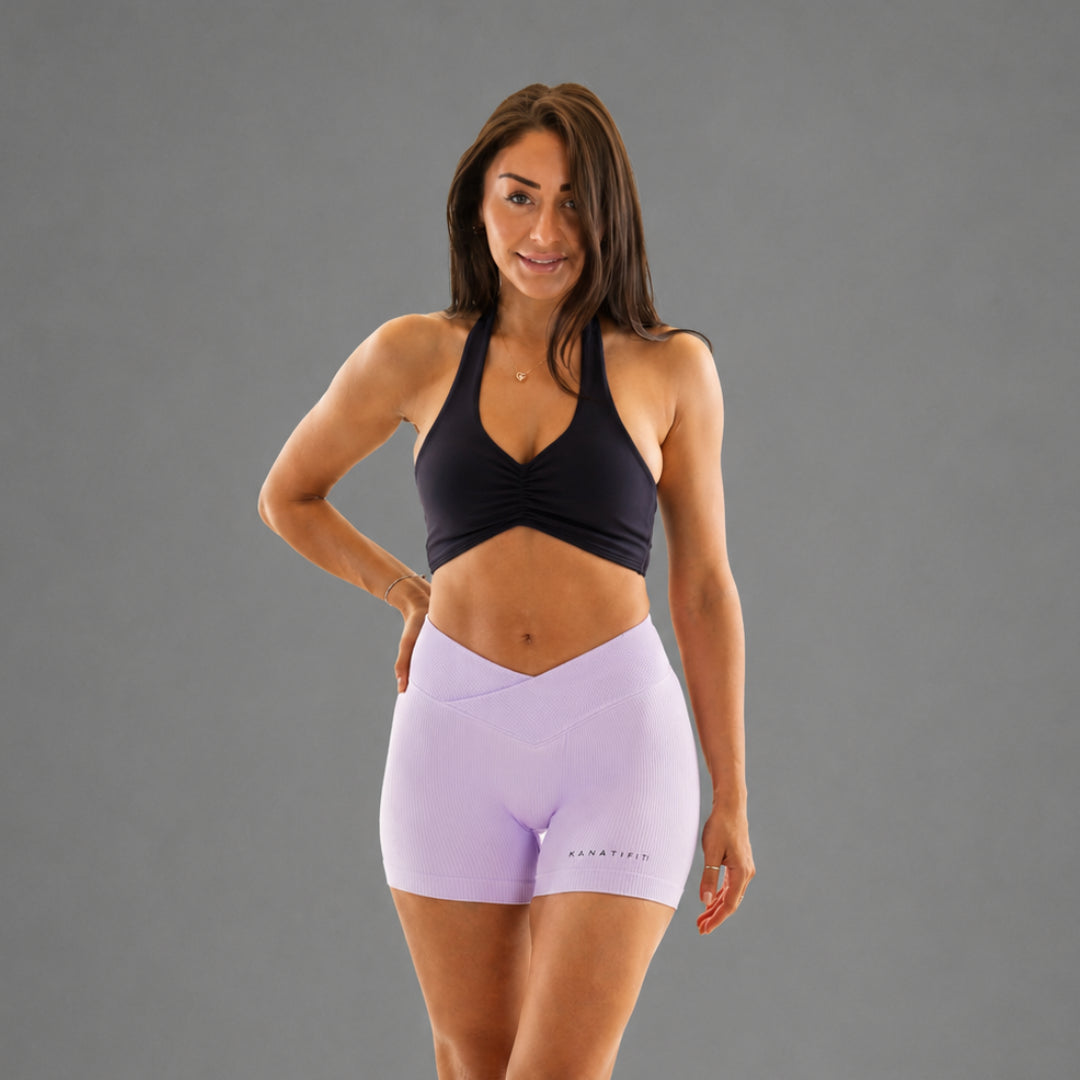 A woman modeling high-waisted light purple athletic shorts and a black sports bra against a solid gray studio background.
