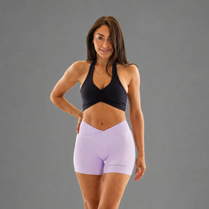 A woman modeling high-waisted light purple athletic shorts and a black sports bra against a solid gray studio background.