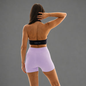 A woman shown from the back wearing light lavender high-waisted athletic shorts and a black sports bra with white text, posed against a solid gray studio background.
