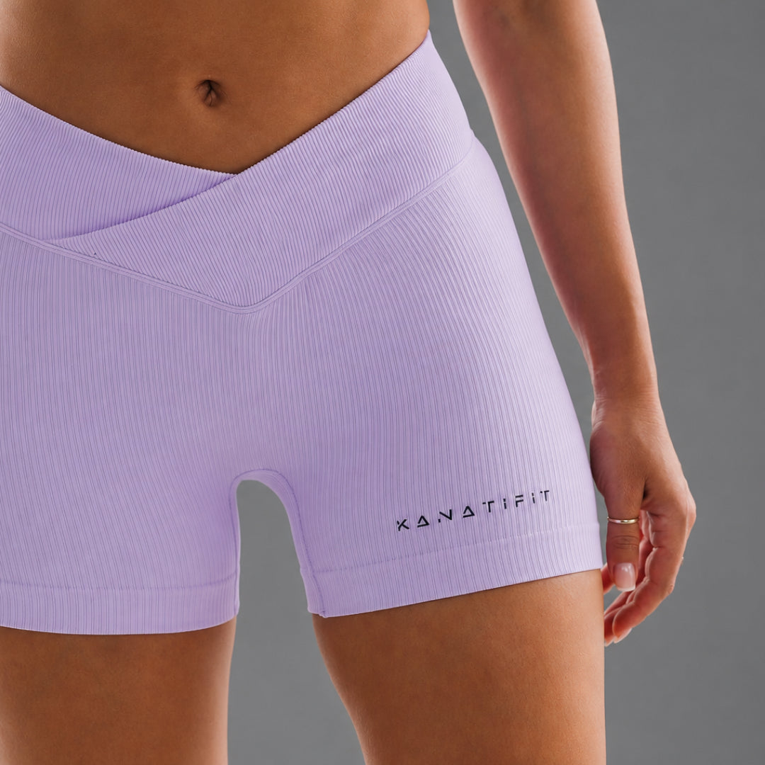 A close-up of a model wearing a grape coloured ribbed athletic shorts with a crossover waistband and the brand name KANATIFIT printed in black near the hem.
