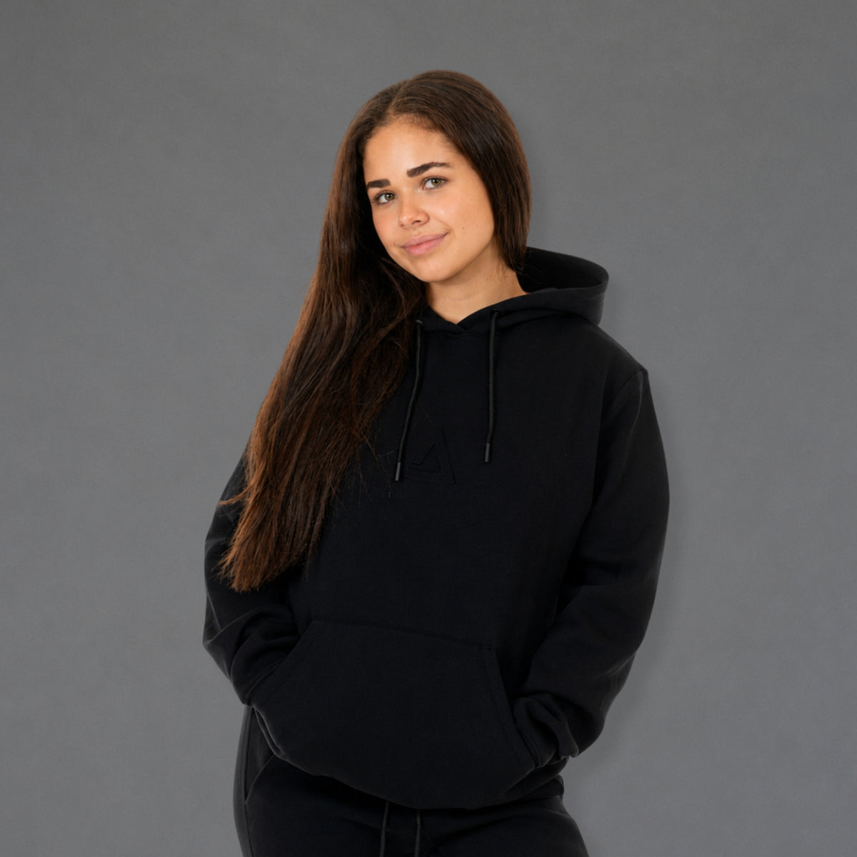 A woman with long dark brown hair wearing a black pullover hoodie with a drawstring hood and kangaroo pockets, standing against a solid gray studio background.