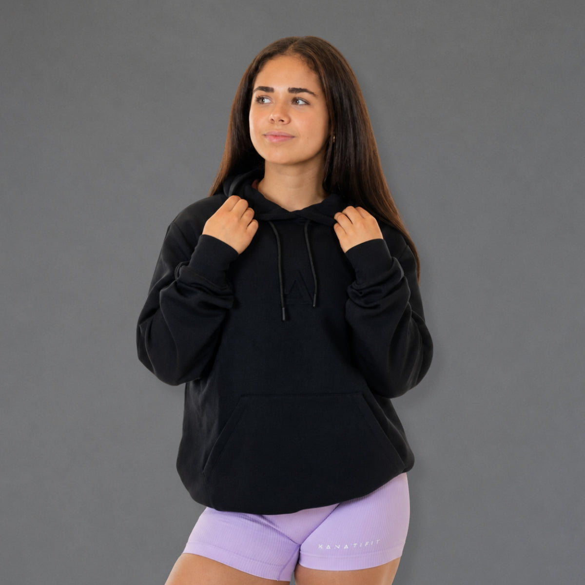 A woman wearing a black pullover hoodie with drawstrings and a front kangaroo pocket, paired with light purple fitted shorts, standing against a solid gray studio background.