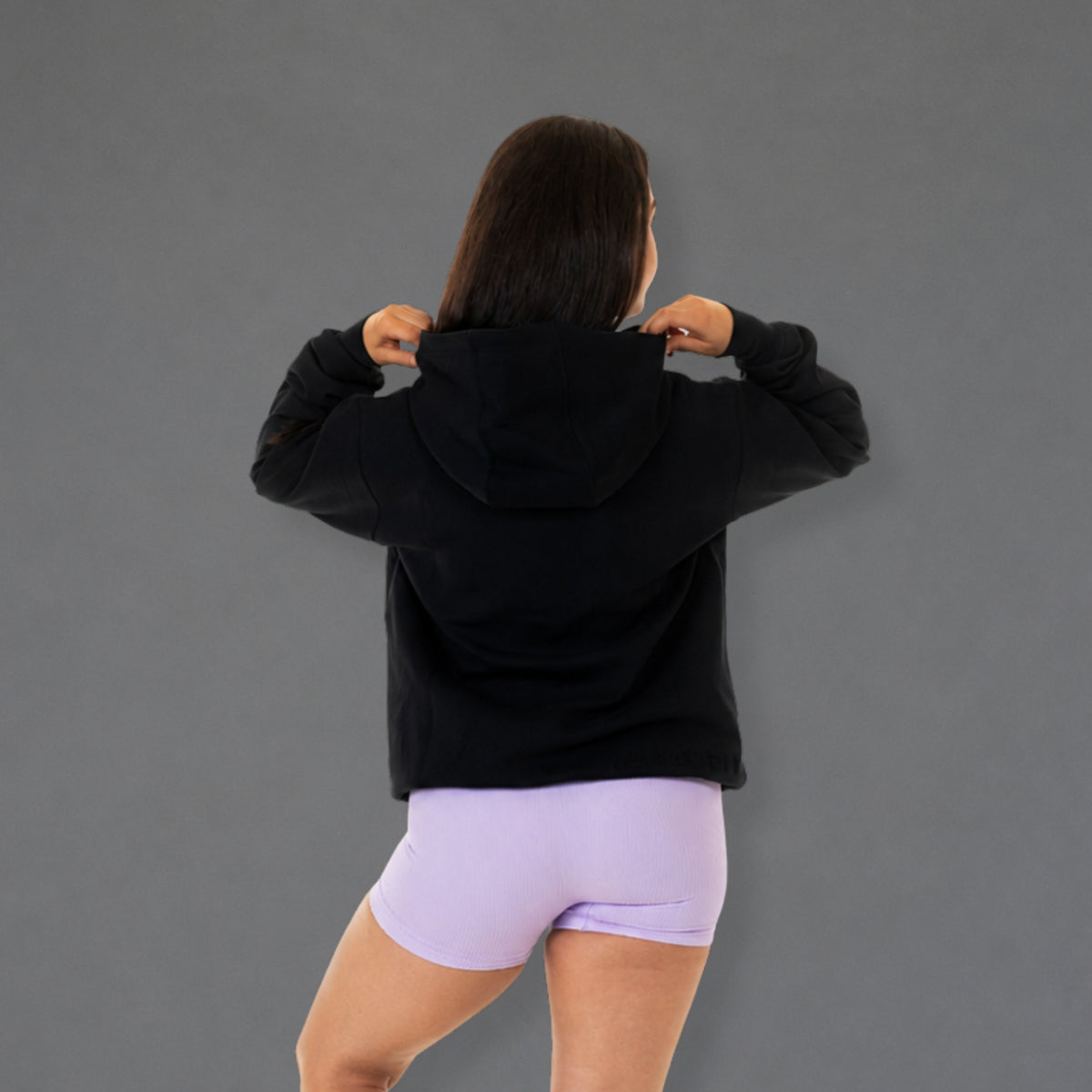 A woman viewed from behind wearing a black cropped hoodie and light purple athletic shorts, posing against a solid gray studio background.