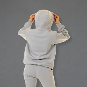 A person shown from the back wearing a light gray hooded sweatshirt and matching jogger pants against a solid gray studio background.