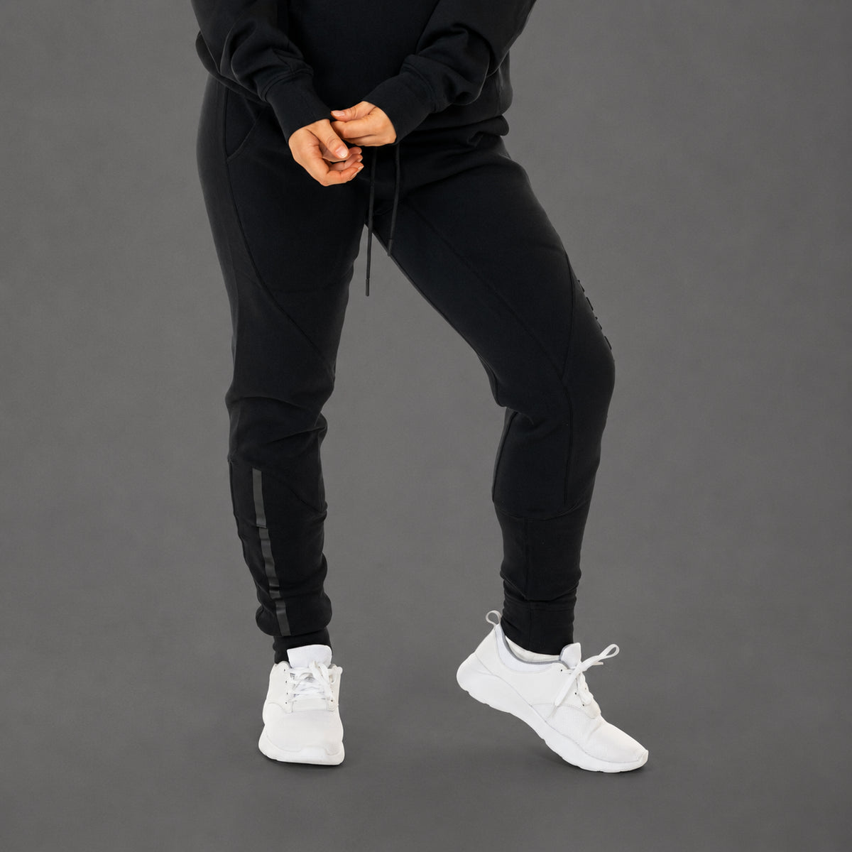 Black jogger sweatpants worn by a model, paired with a black hoodie and white sneakers, shown from the waist down against a solid gray studio background.