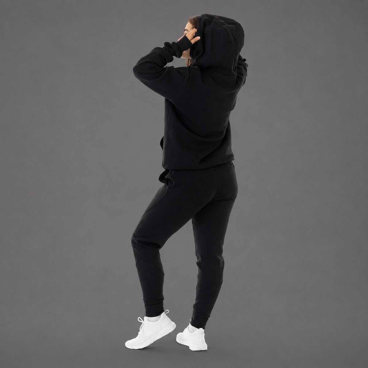 A model wearing a matching black hoodie and jogger sweatpants set, shown from the back and side against a solid gray studio background.