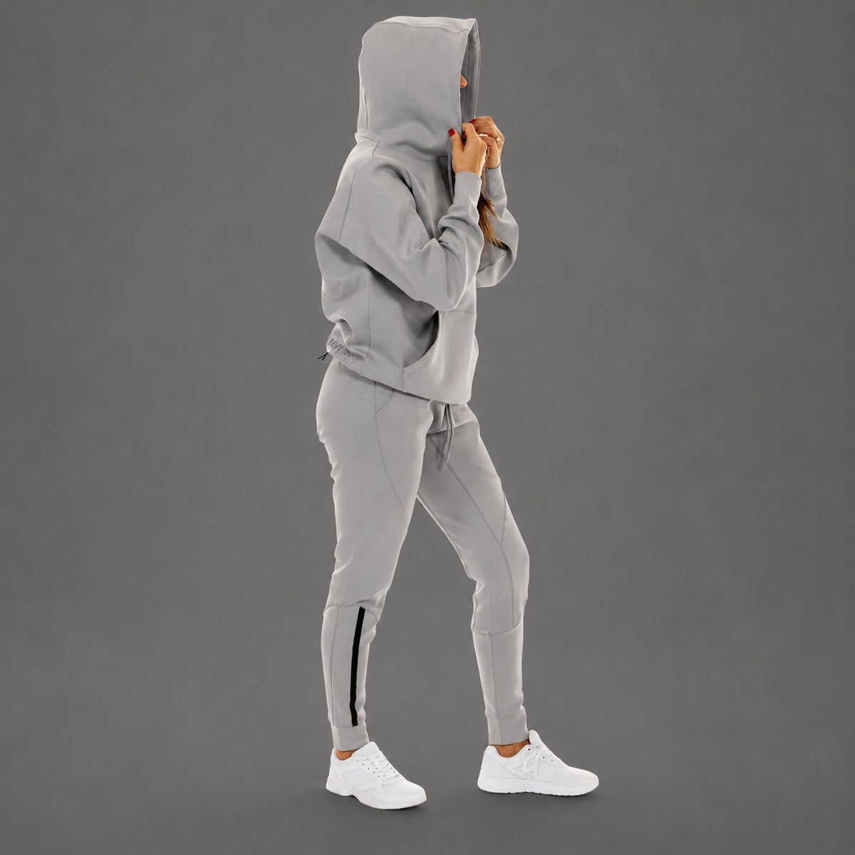 A model wearing a light gray hooded sweatshirt and matching jogger pants with a black side stripe, paired with white sneakers, shown against a solid gray studio background.