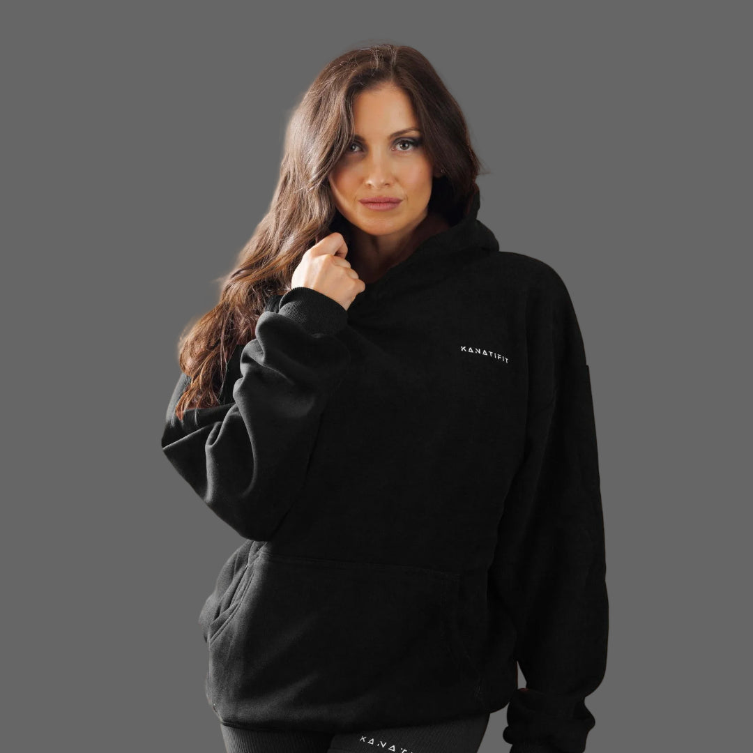A woman with long brown hair wearing a black pullover hoodie with a small white 'KANITFIT' logo on the left chest, posed against a solid dark gray studio background.