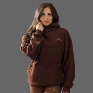 A woman wearing a dark brown pullover hoodie with a small white 'KANATIFIT' logo on the left chest, posed against a solid dark gray studio background.
