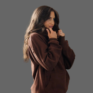 A woman with long wavy brown hair wearing a brown fleece pullover hoodie, posing against a solid dark gray background.